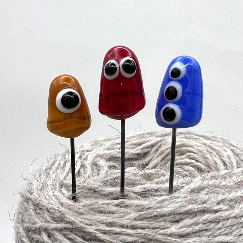 Three little glass monster pins, one red, one yellow, one blue. All three have googly glass eyes. Pins are stuck into a ball of yarn.