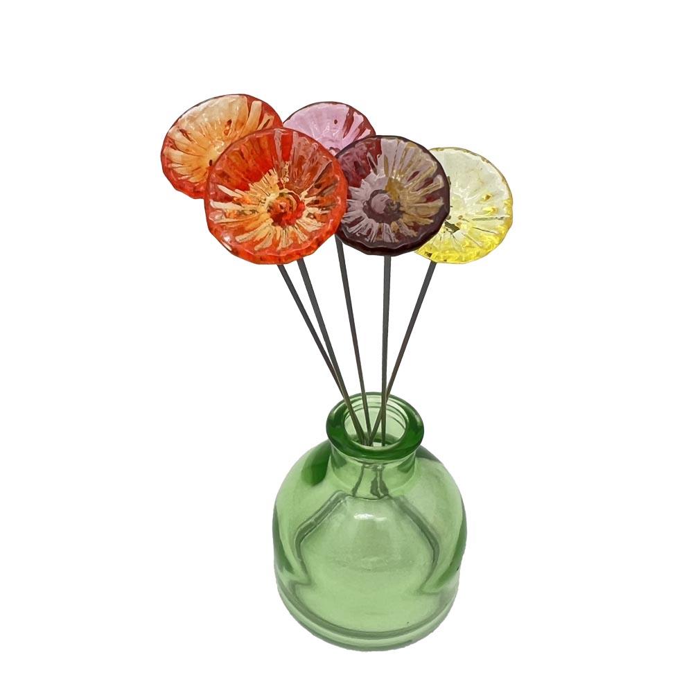 five little glass flowers made with transparent glass on long steel stems sit in a green vase.
