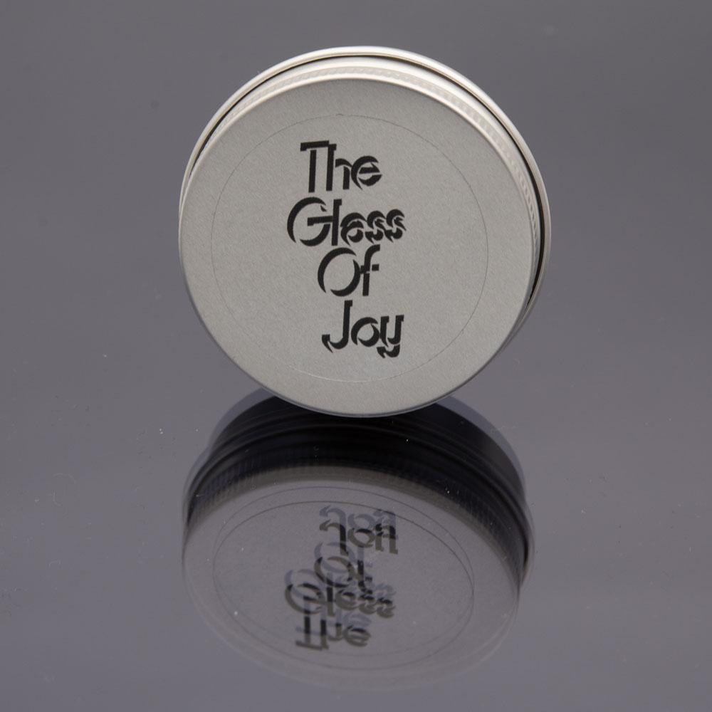 Small round metal tin photographed on a black reflective surface. The lid has a sticker with The Glass Of Joy logo.
