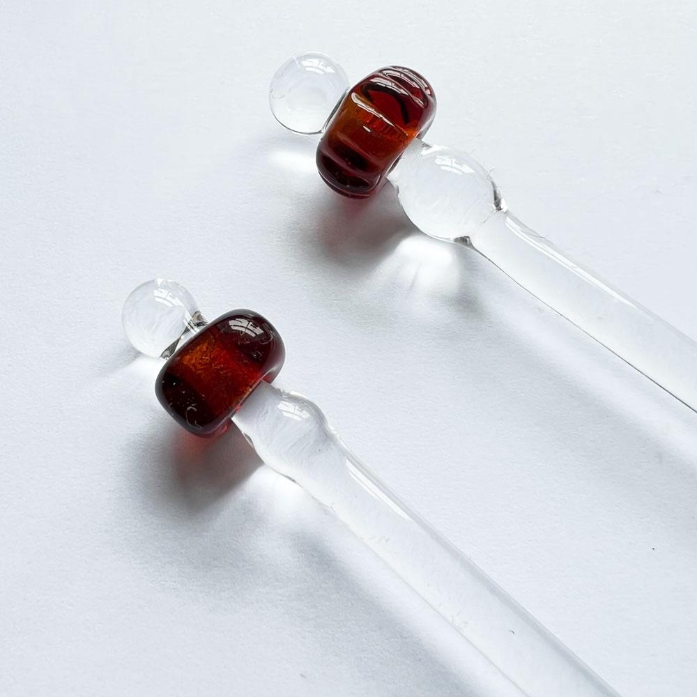 Close up of two cocktail stirrers each with a viking style bead made from amber glass. Both beads are simple donut shaped. The stirrers are made from clear glass.