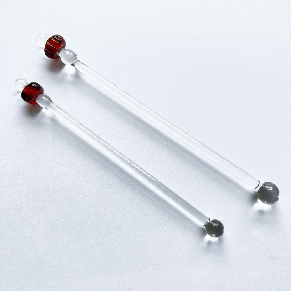 Two cocktail stirrers each with a viking style bead made from amber glass. Both beads are simple donut shaped. The stirrers are made from clear glass.