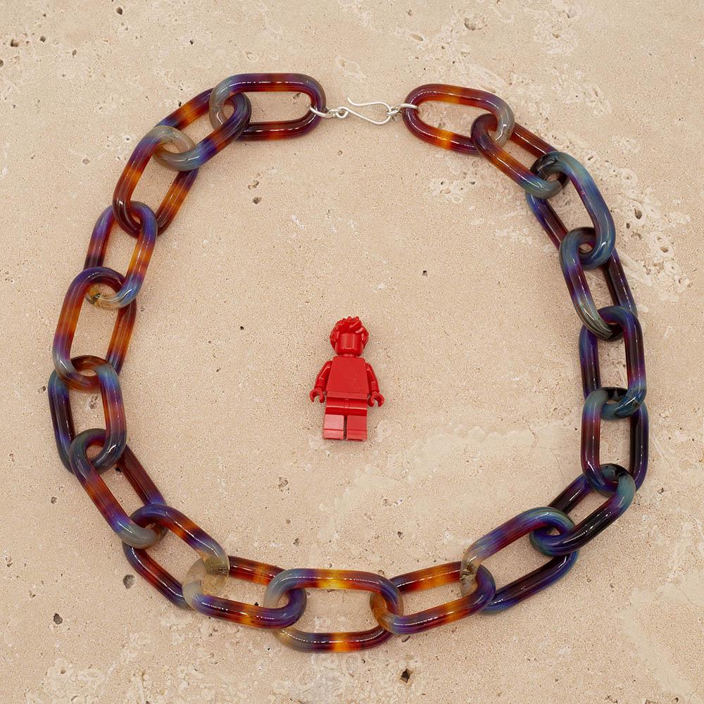 Transparent multicolour glass chain link necklace with a sterling silver hook and loop catch. Each link has blue, yellow, orange, purple and pink. The necklace sits on a sandstone tile and is shown with a red lego figure for scale.
