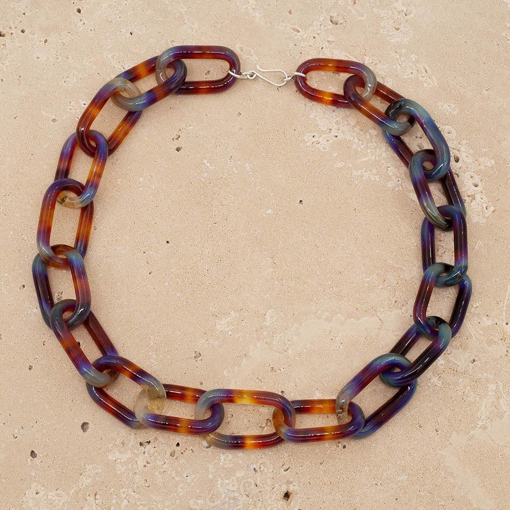 Transparent multicolour glass chain link necklace with a sterling silver hook and loop catch. Each link has blue, yellow, orange, purple and pink. The necklace sits on a sandstone tile.