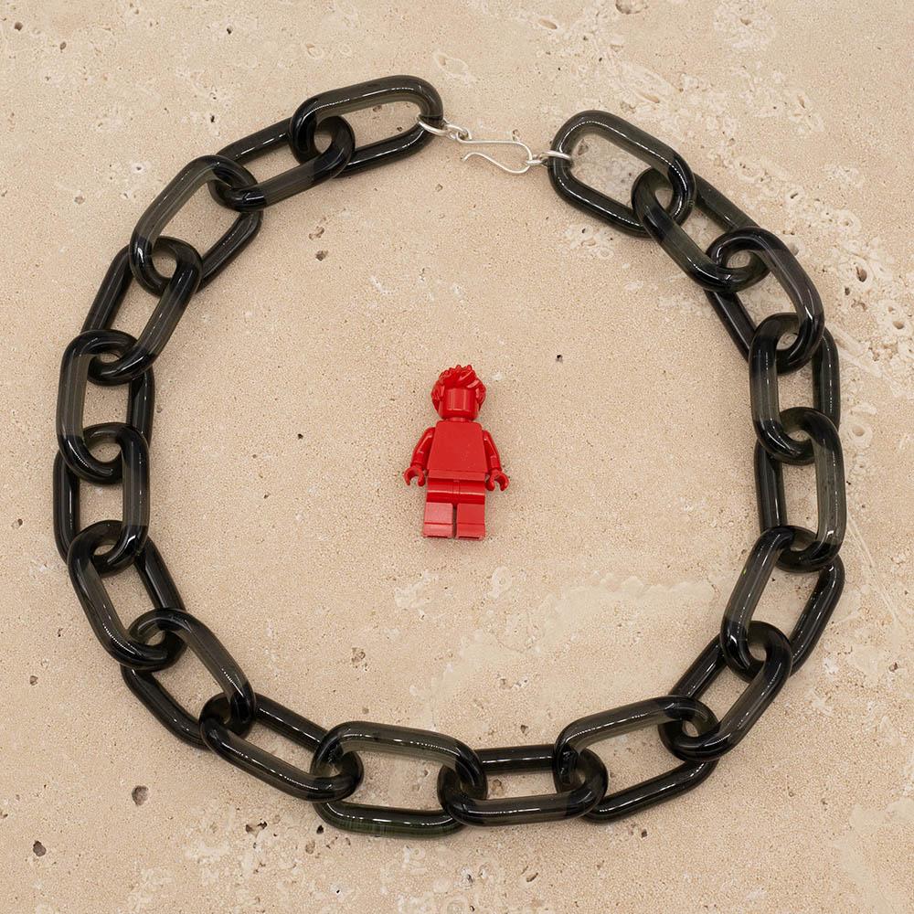 Transparent black glass chain link necklace with a sterling silver hook and loop catch. The necklace sits on a sandstone tile, Shown with a red lego figure for scale.
