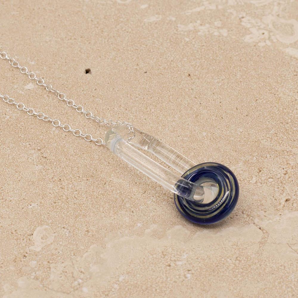 Close up of a clear glass link which passes through a beads made aurora trails glass, giving spirals of blue and clear. The link hangs from a silver chain. Shown on a sandstone background.
