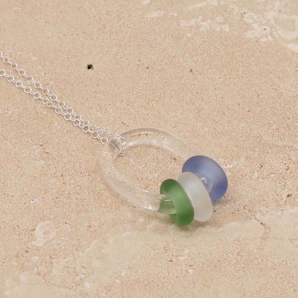 Close up of a clear glass link which passes through 3 frosted beads made from green, clear and blue glass. The link hangs from a silver chain. Shown on a sandstone background.