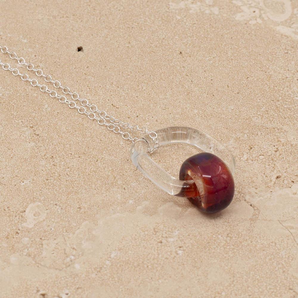 Close up of a clear glass link which passes through a barrel shaped bead made with pink mai tai glass. The link hangs from a silver chain. Shown on a sandstone background.