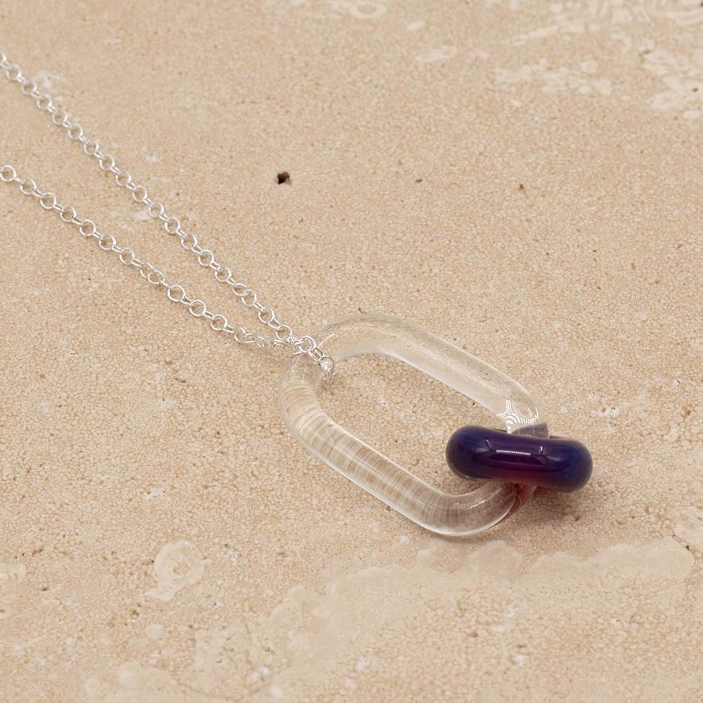 Close up of a clear glass link which passes through a bead made with purple and blue mai tai glass. The link hangs from a silver chain. Shown on a sandstone background.