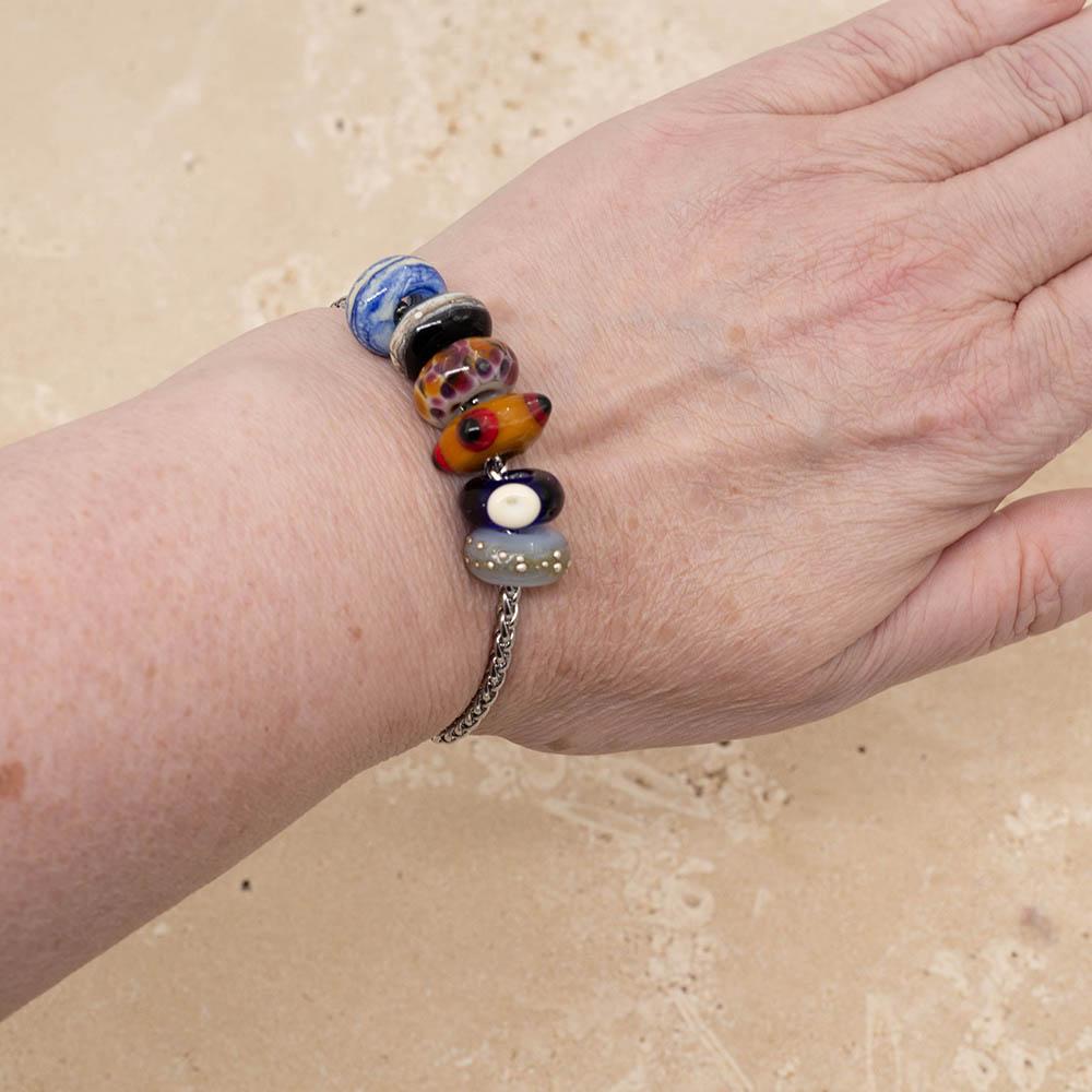 6 big hole beads on a stainless steel chain bracelet, shown worn on a wrist. The beads are swirled blue and ivory, black with a band of ivory, pale grey with speckles of orange and pink, gold with raised red and black dots, dark blue with a clear dot and pale blue with dots of fine silver.