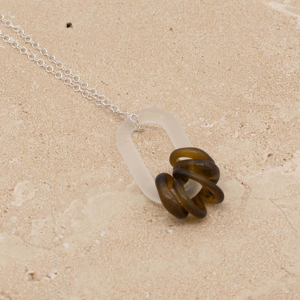 Close up of a frosted glass link which passes through 5 beads made from a brown glass marmite jar. The link hangs from a silver chain. Shown on a sandstone background.