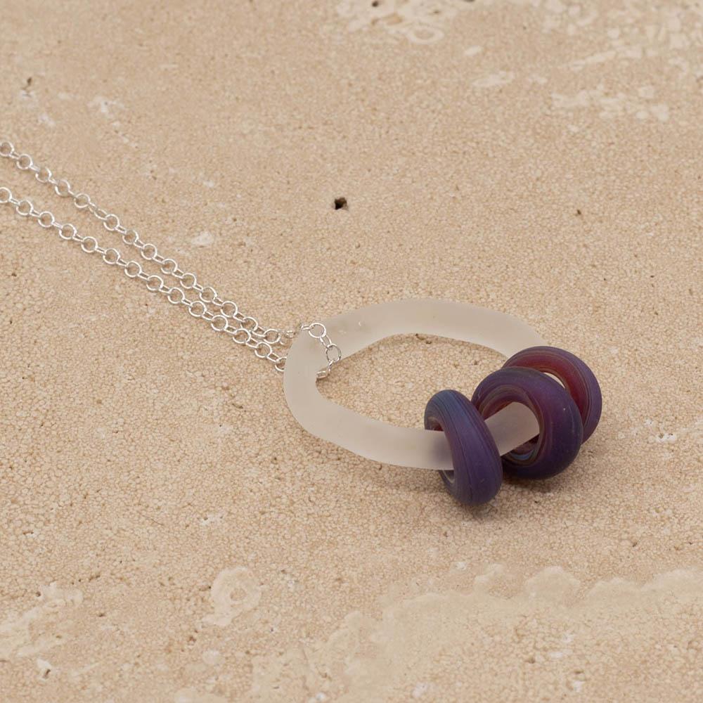 Close up of a frosted glass link which passes through 3 frosted blueberry coloured beads. The link hangs from a silver chain. Shown on a sandstone background.