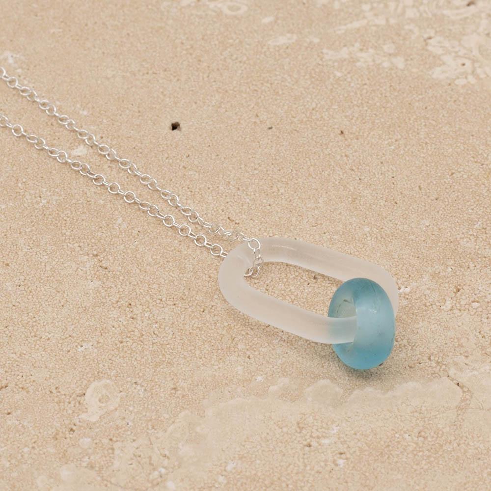 Close up of a frosted glass link which passes through a bead made from a turquoise Bombay Sapphire gin bottle. The link hangs from a silver chain. Shown on a sandstone background.