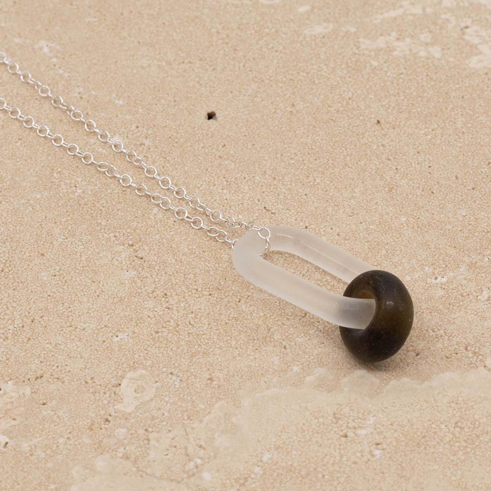 Close up of a frosted glass link which passes through a bead made from a brown marmite jar. The link hangs from a silver chain. Shown on a sandstone background.