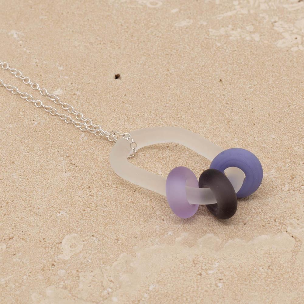 Close up of a frosted glass link which passes through 3 beads made from different shades of purple glass The link hangs from a silver chain. Shown on a sandstone background.