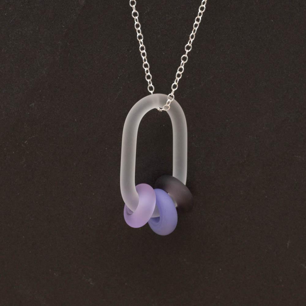 Close up of a frosted glass link which passes through 3 beads made from different shades of purple glass The link hangs from a silver chain. Shown on a slate background.