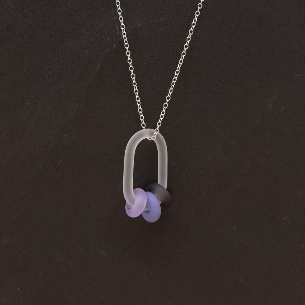 A frosted glass link which passes through 3 beads made from different shades of purple glass The link hangs from a silver chain. Shown on a slate background.