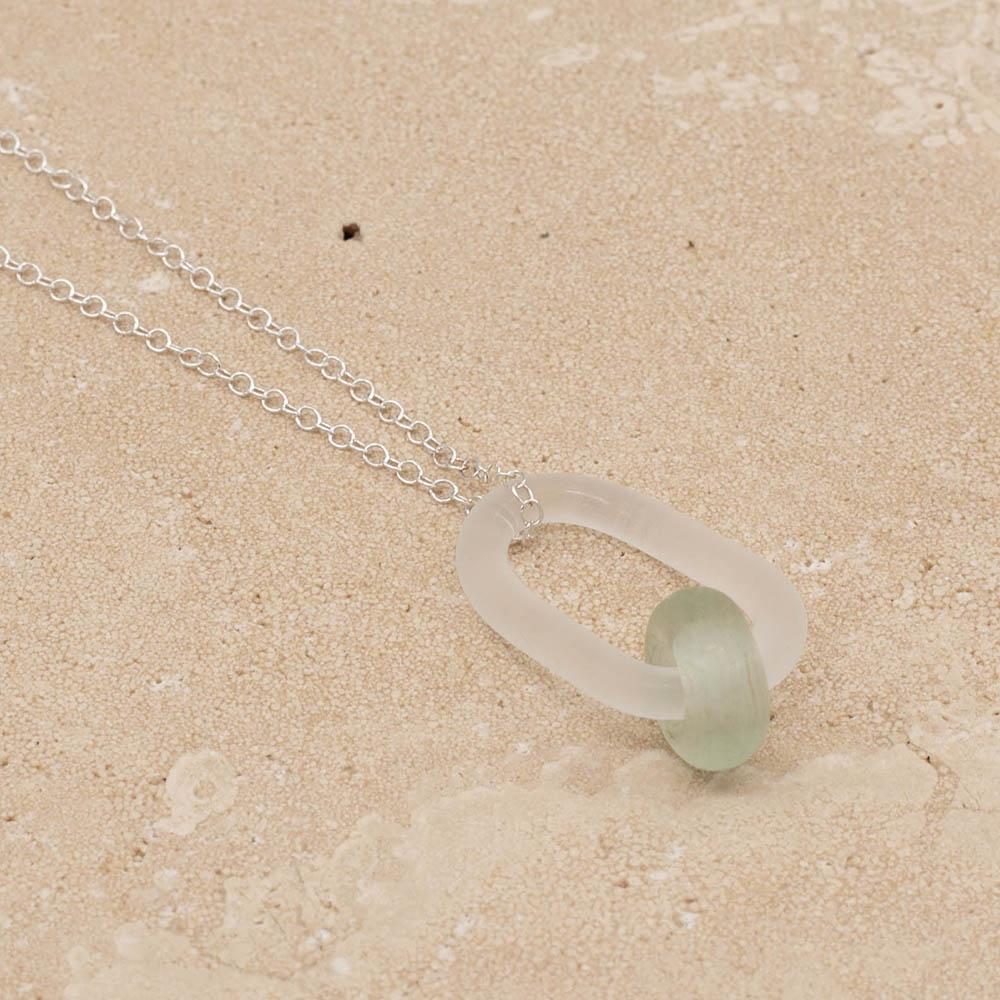 Close up of frosted glass link which passes through a bead made from pale green wine bottle glass. The link hangs from a silver chain. Shown sitting on a sandstone tile.