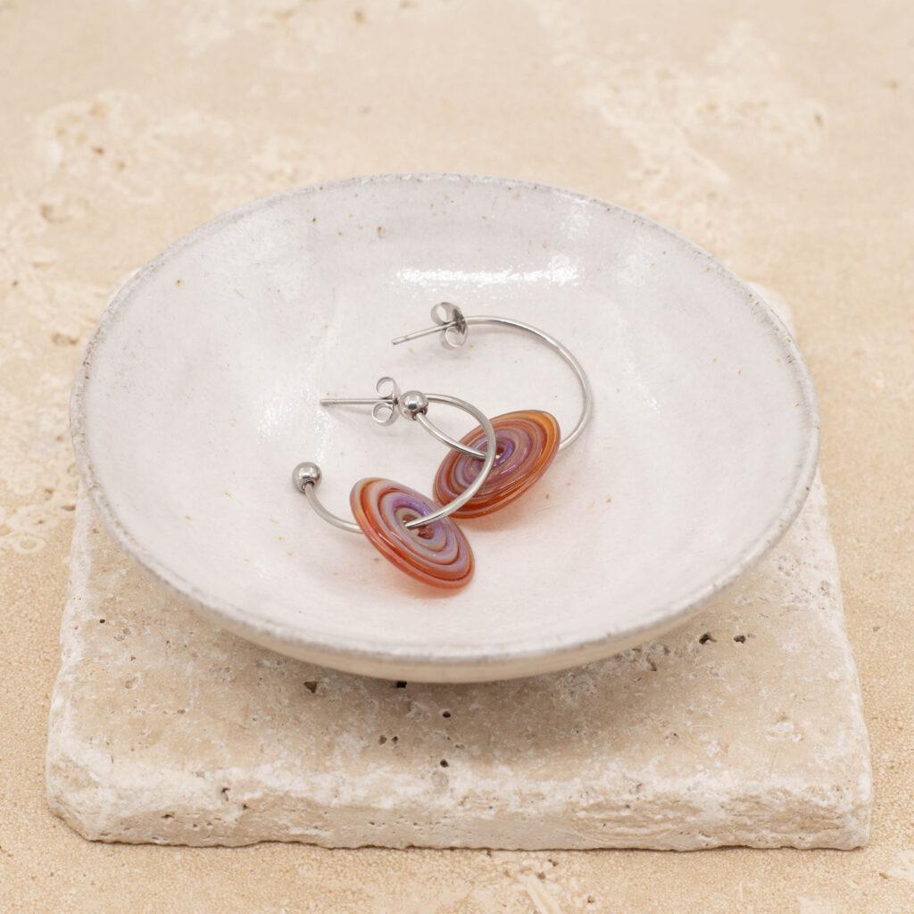 A pair of hoop earrings with amber purple glass discs sitting in a small white bowl.