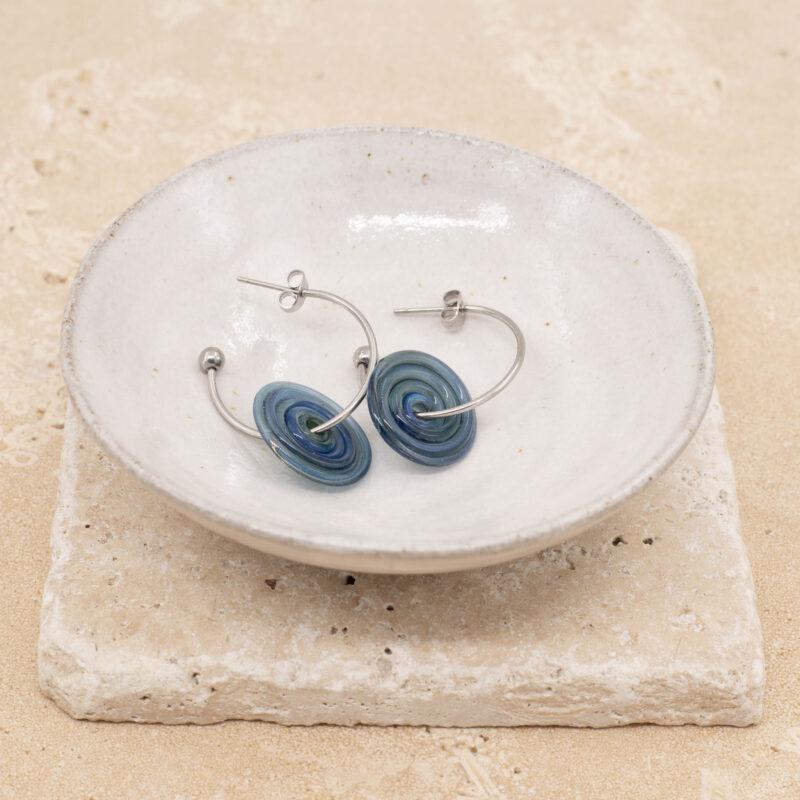 A pair of hoop earrings with blue and green glass discs sitting in a small white bowl.