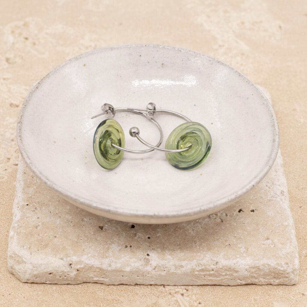 A pair of hoop earrings with transparent light green glass discs sitting in a small white bowl.