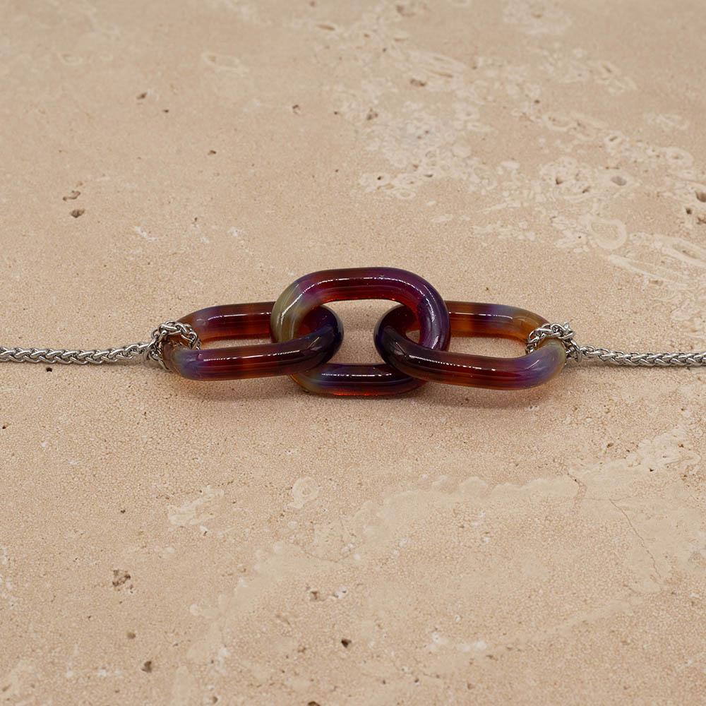 Close up of necklace with three mai tai glass links giving purple, blue and amber. The links are fastened to a chain to make a necklace. Shown on a sandstone background.