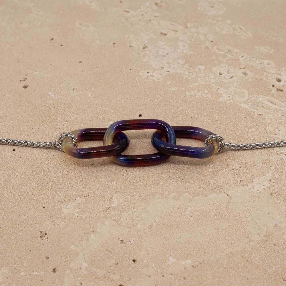 Close up of necklace with three mai tai glass links giving purple, blue and yellow. The links are fastened to a chain to make a necklace. Shown on a sandstone background.