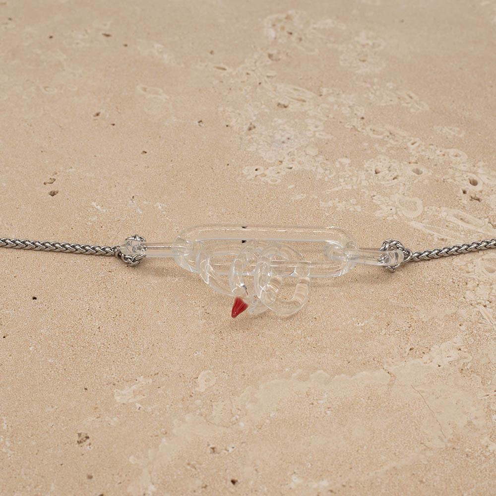 Close up of necklace with long clear glass link and five smaller clear links. One smaller link has a red accent. Shown on a sandstone background.