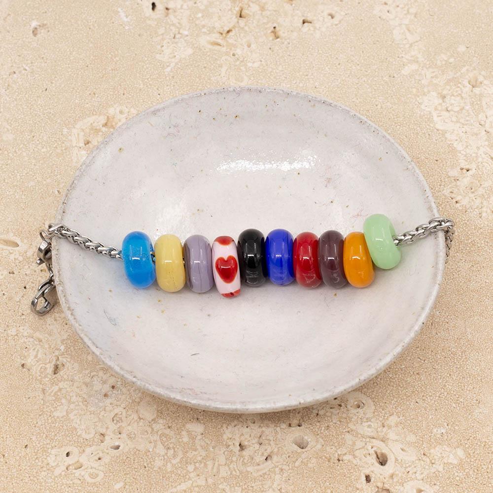 10 beads representing the Taylor Swift Eras Tour on a stainless steel chain bracelet sitting in a small white bowl. The colours are pale green, gold, deep purple, red, bright blue, black, pale pink with red hearts, lilac, pale yellow and bright turquoise.