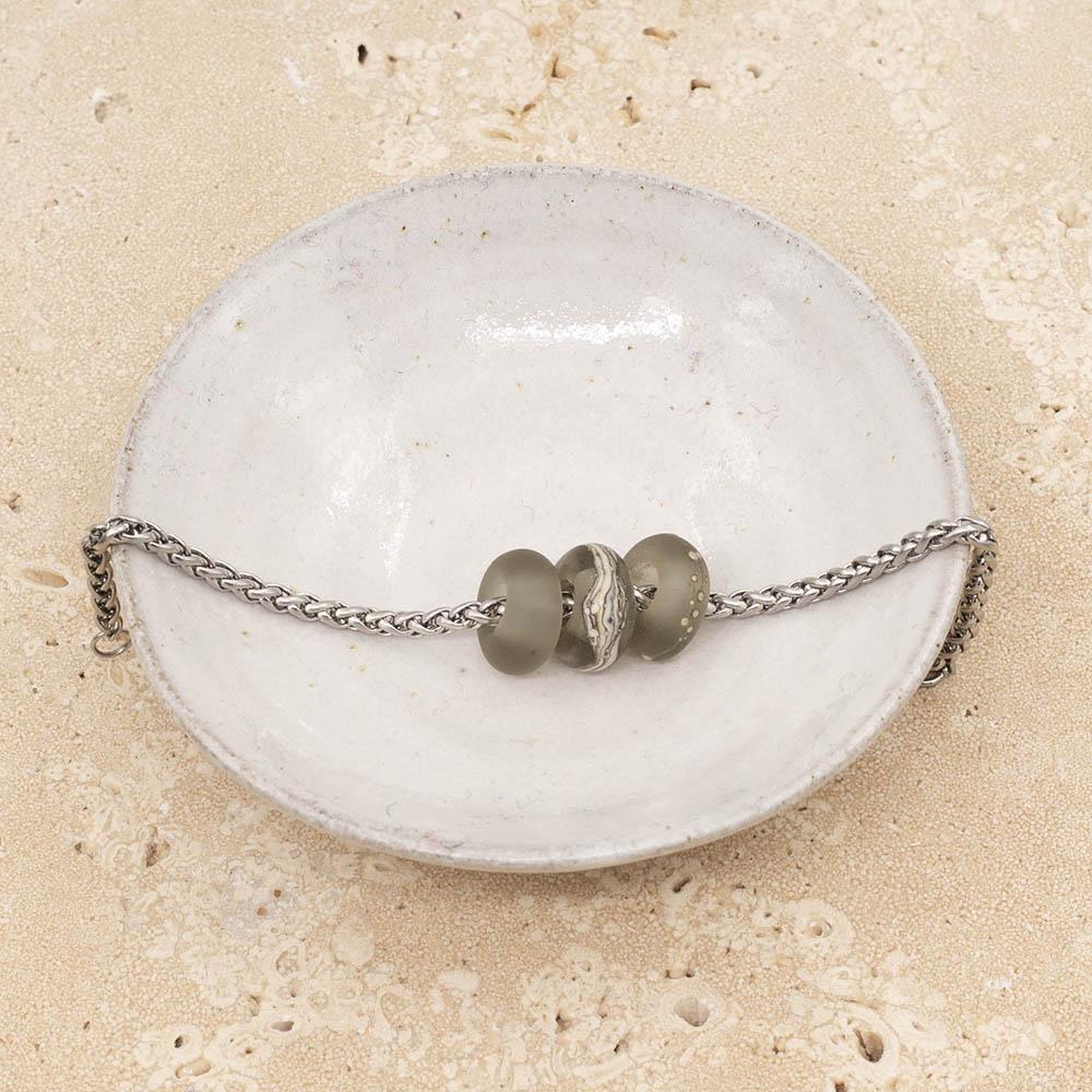 3 frosted grey glass beads with big holes on a stainless steel chain bracelet sitting in a small white bowl.
