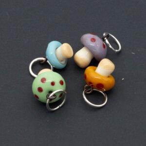 Mushroom charms. The mushrooms are golden yellow, green.turquoise and purple and all four have red dots. Each mushroom has a metal hanging loop.