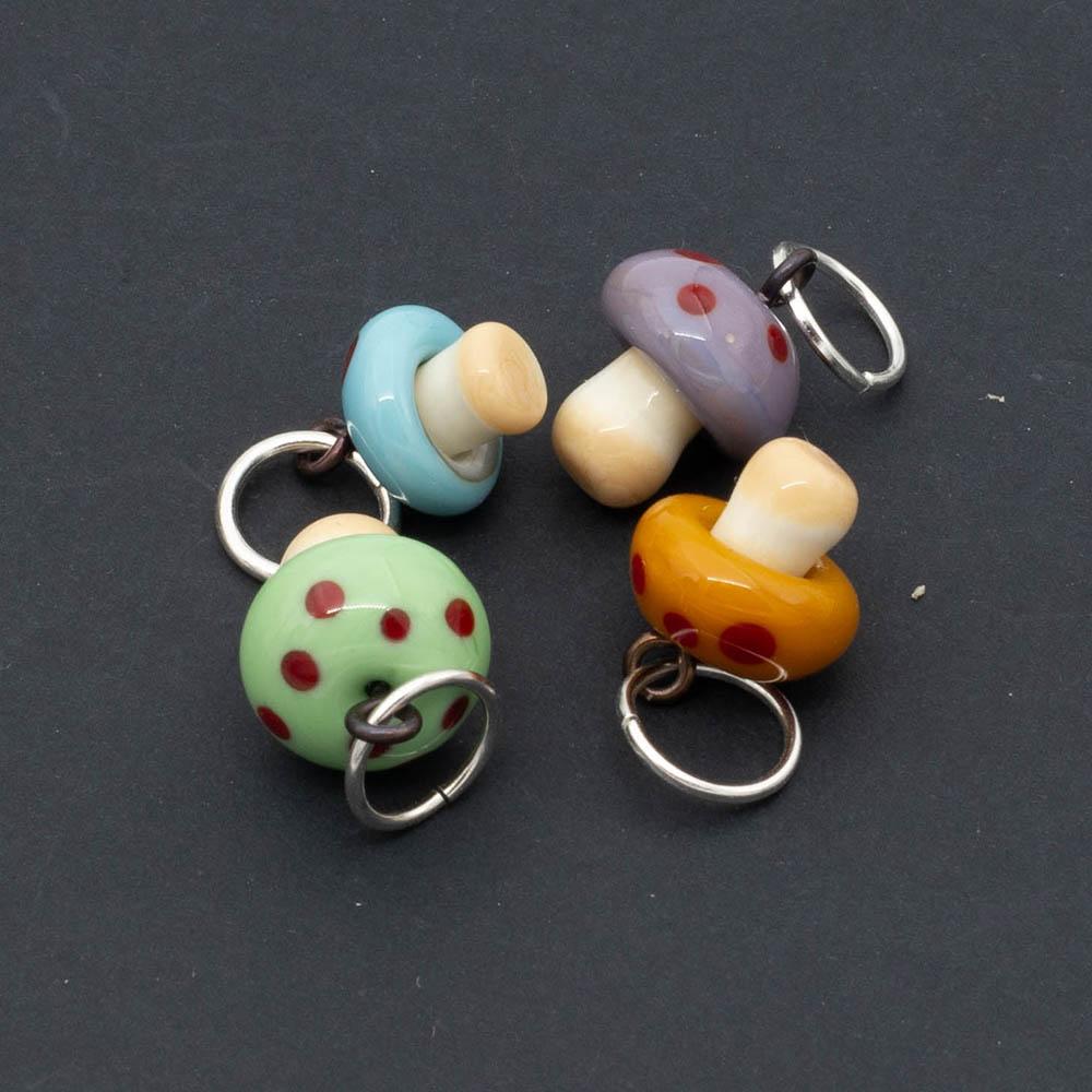 Mushroom charms. The mushrooms are golden yellow, green.turquoise and purple and all four have red dots. Each mushroom has a metal hanging loop.