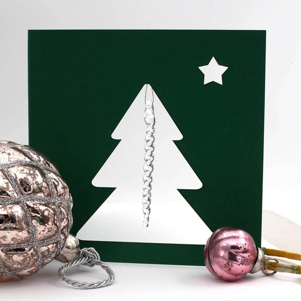 Front of dark green greetings card. The card is square. The card has a cutout of a Christmas tree and small star. A clear glass icicle hangs from the top of the tree. Styled with two pink glass baubles.