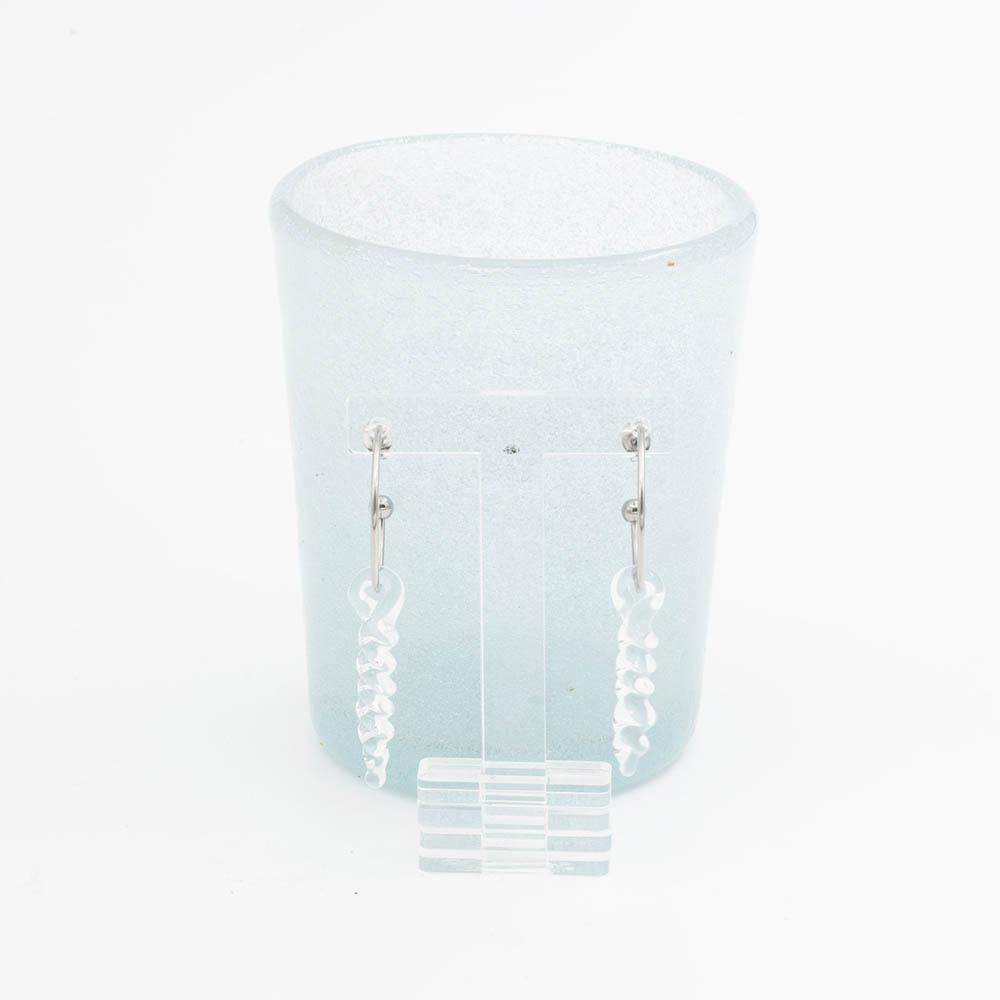 two large clear icicle glass icicles on earring hoops hang on a clear stand in front of a pale blue tumbler